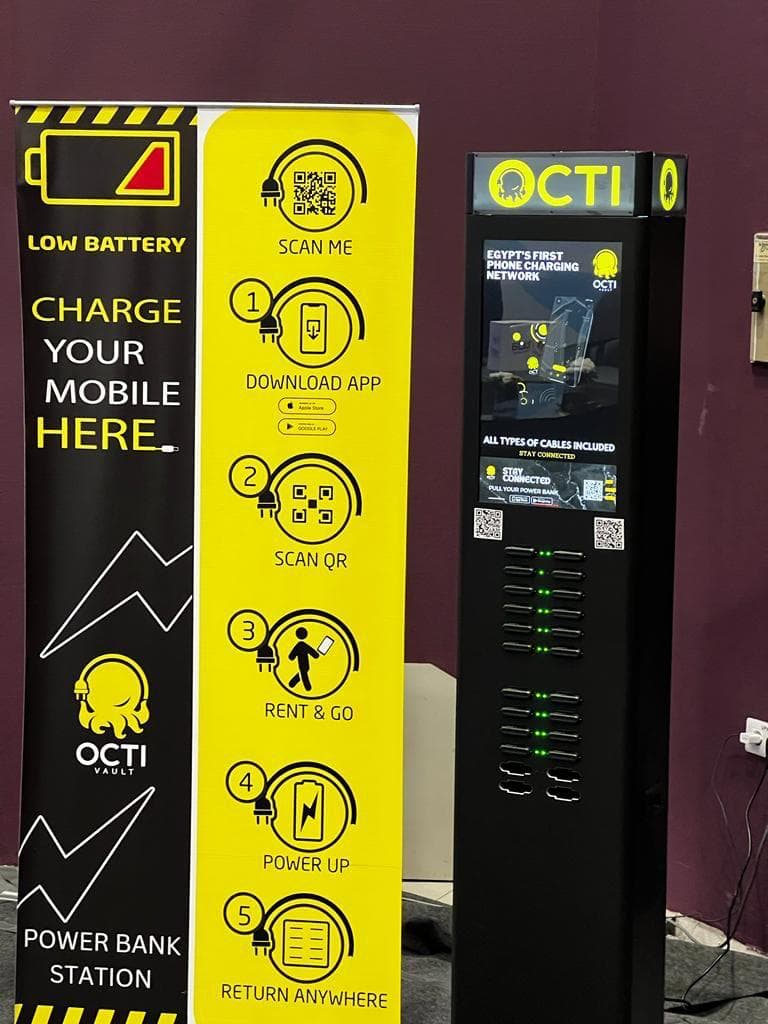 A charging station indoors with its lit screen and printed wayfinding panel facing the walkway